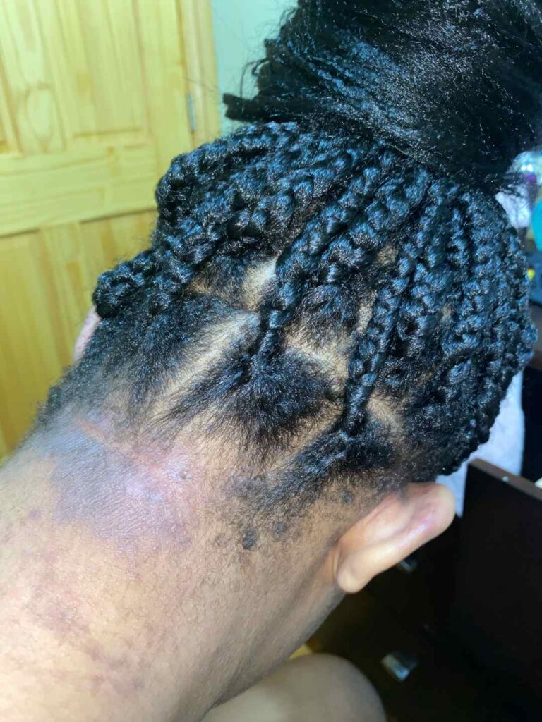 A close-up of the back of a person’s head shows neatly done box braids gathered into a bun, with the nape of the neck and some natural hair visible. The background includes a wooden door and part of a chair.