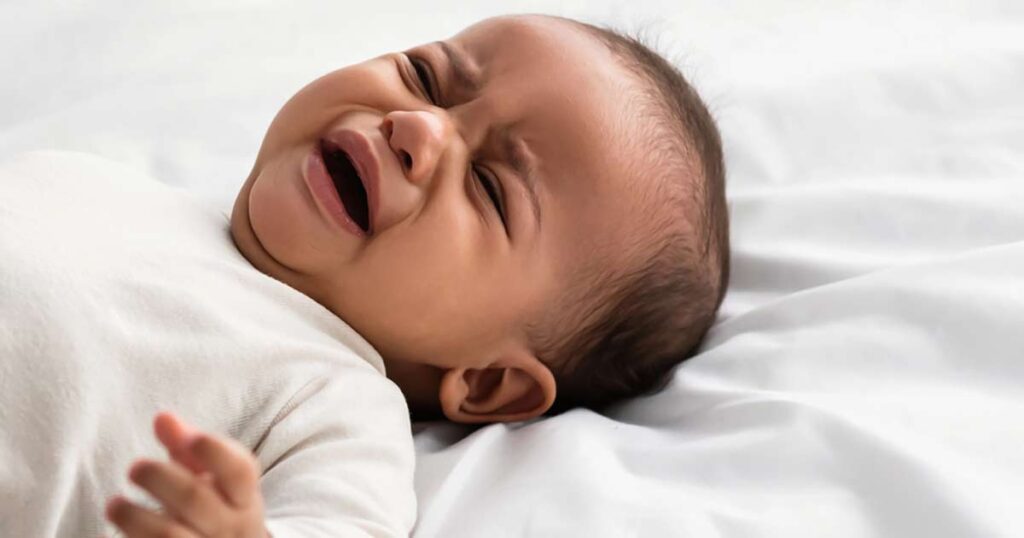 A baby lying on a white blanket is crying, with eyes closed, mouth open, and hand reaching out.