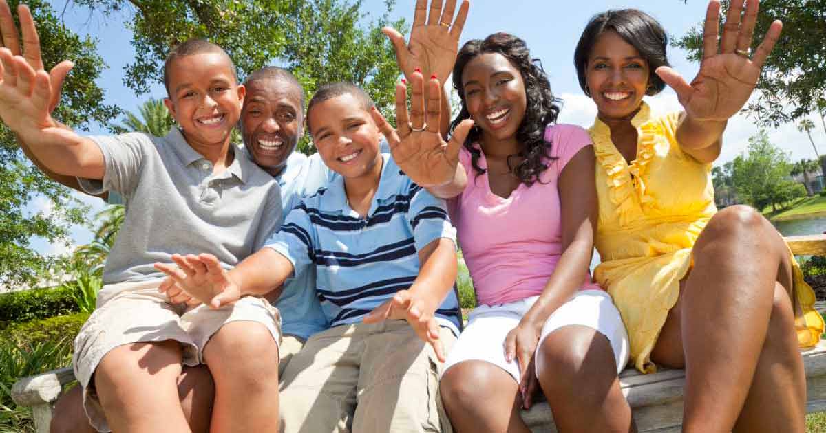 A smiling family of five sits outdoors on a bench, waving at the camera. The group includes two adults and three children, with trees and greenery in the background on a sunny day.