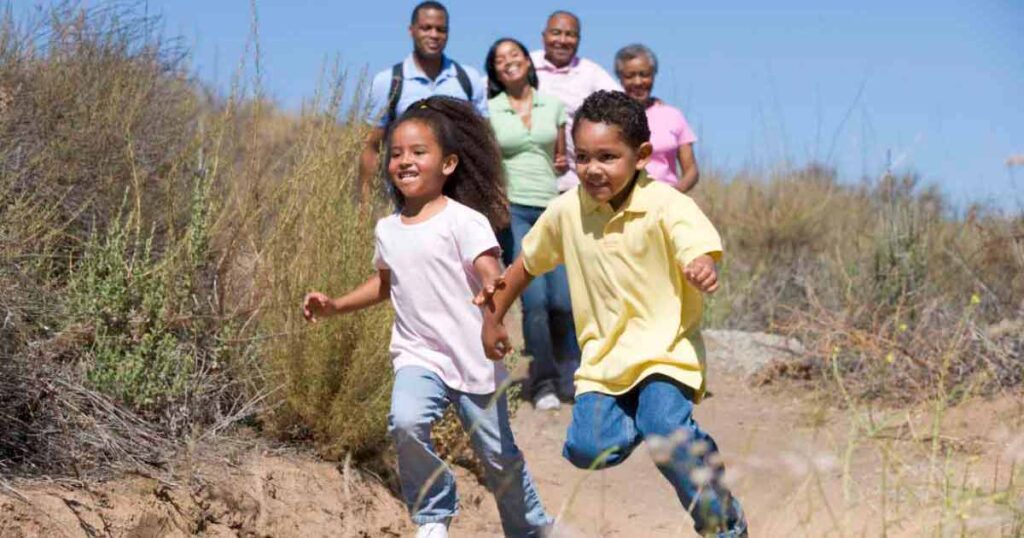 Two young children run along a dirt trail in a grassy outdoor setting, smiling and holding hands. Four adults walk behind them, also smiling, under a clear blue sky.