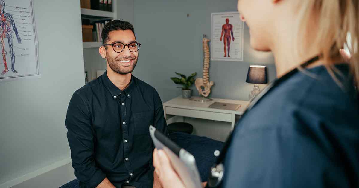 A smiling man wearing glasses sits on an exam table in a medical office, facing a healthcare professional holding a tablet. Medical charts and a model spine are visible in the background.