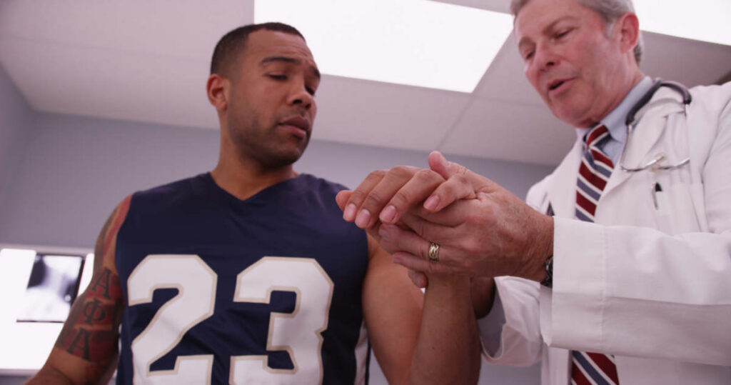A male doctor examines the hand of a male athlete wearing a navy jersey with the number 23 in a medical office. The athlete looks concerned as the doctor inspects his hand.