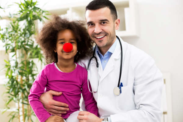 A smiling doctor with a stethoscope sits beside a young girl wearing a red clown nose and pink shirt. They both look at the camera in a cheerful, brightly lit room with a plant in the background.