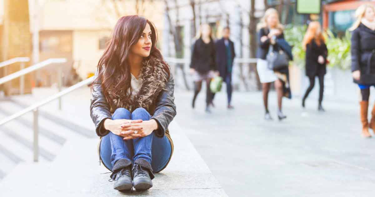 A woman with long dark hair sits on a ledge outdoors, wearing a black jacket with a fur collar and blue jeans, while people walk by in the blurred background.