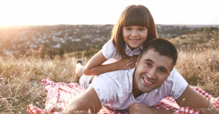 A man and a young girl smile while lying on a red and white checkered blanket in a grassy field at sunset. The girl is playfully lying on the man's back, both looking happy.