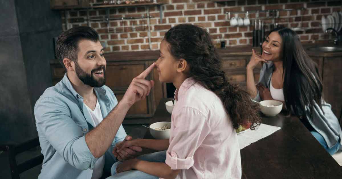 A man playfully touches a young girl's nose as they smile at each other, while a woman in the background laughs. They are sitting together at a kitchen table with bowls of food.