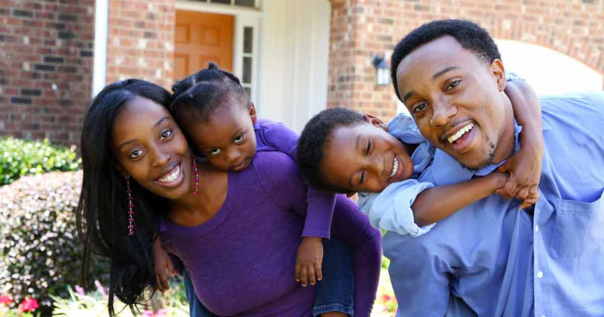 A smiling family of four poses outside their home. The mother holds a young girl on her back, while the father holds a young boy on his back. They all look happy and are dressed in shades of blue and purple.