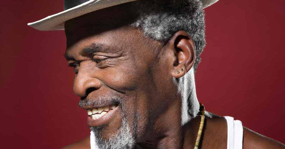 Smiling older man with gray hair and beard wearing a white hat, white tank top, and beaded necklace, against a red background.