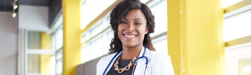 A smiling woman wearing a white medical coat and a stethoscope stands indoors in front of large yellow pillars and windows.