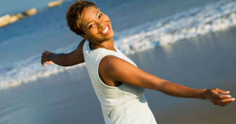 A smiling woman with short hair stands on a beach with her arms outstretched, wearing a sleeveless white top, with waves and blue ocean water in the background.
