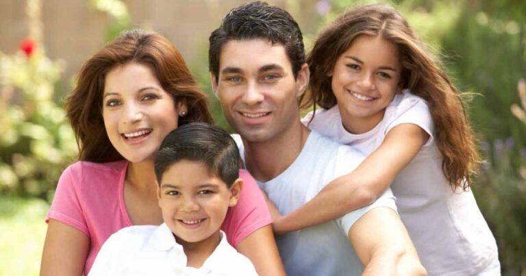 A smiling family of four poses outdoors. The parents sit close together, with a young boy in front and a young girl hugging her father's shoulder from behind. Greenery is visible in the background.