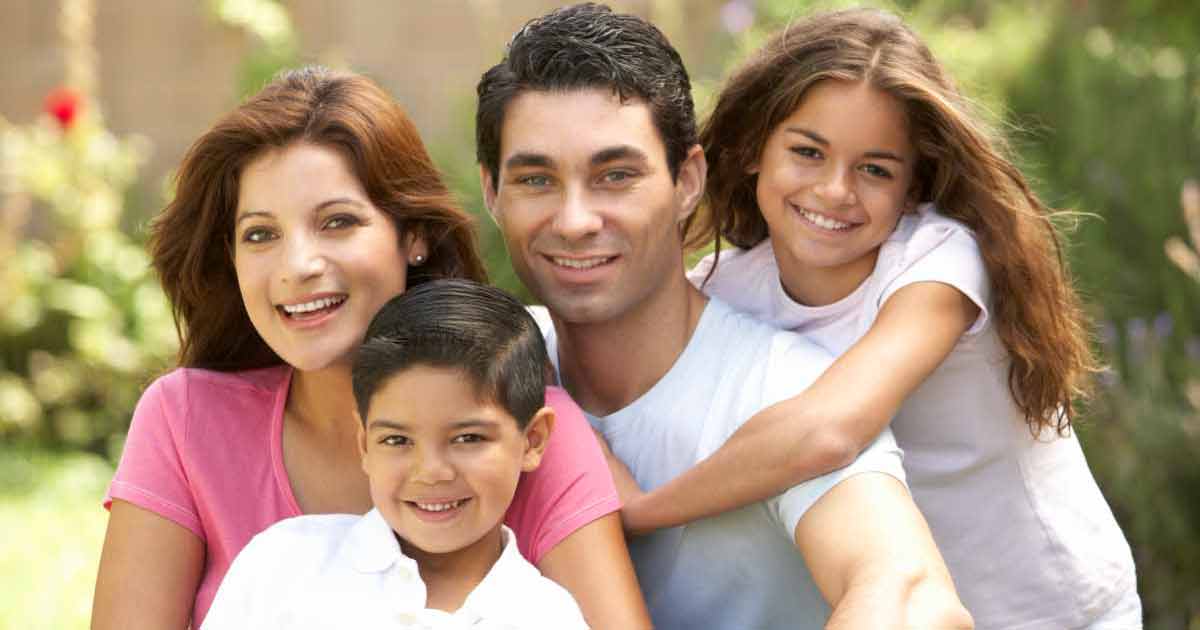 A smiling family of four poses outdoors. The parents sit close together, with a young boy in front and a young girl hugging her father's shoulder from behind. Greenery is visible in the background.