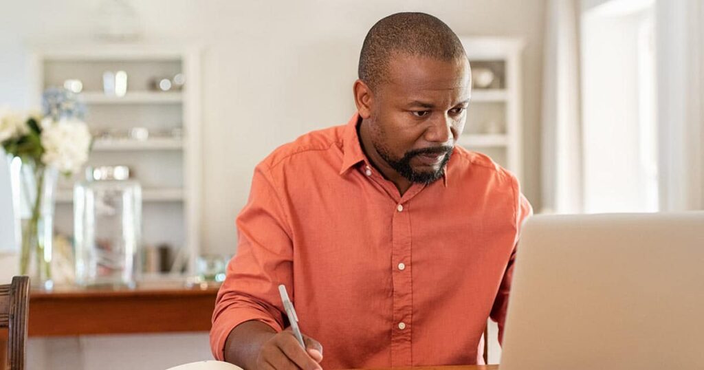 A man in an orange shirt sits at a table, focused on his laptop while writing notes with a pen. The background shows shelves and a vase of flowers in a well-lit room.