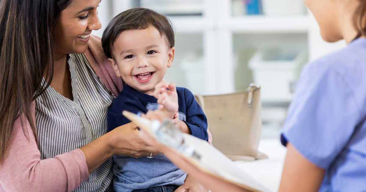 Una mujer sonriente sostiene a un niño pequeño feliz en su regazo mientras un trabajador de la salud con uniforme médico habla con él y sostiene un portapapeles, posiblemente durante una consulta médica.