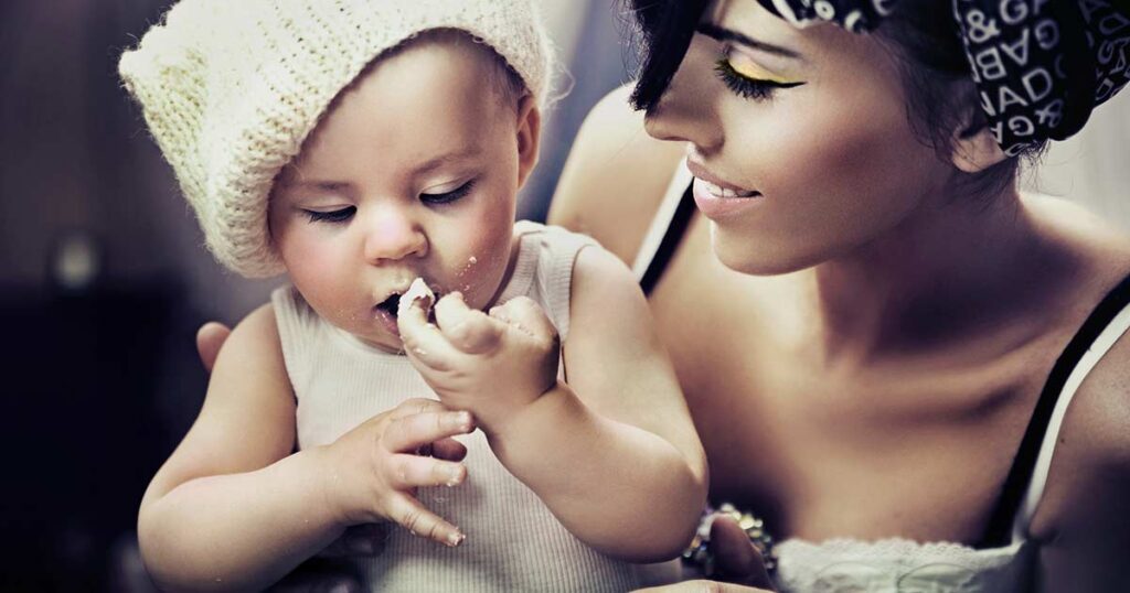 A smiling woman watches as a baby in a white knit hat and tank top eats with their hands, crumbs on their face and fingers. The scene is warm and intimate, suggesting a caring relationship.
