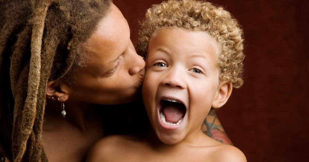 An adult with dreadlocks kisses a young, curly-haired child on the cheek. The child has an open mouth and wide eyes, appearing surprised or excited. Both are against a dark red background.