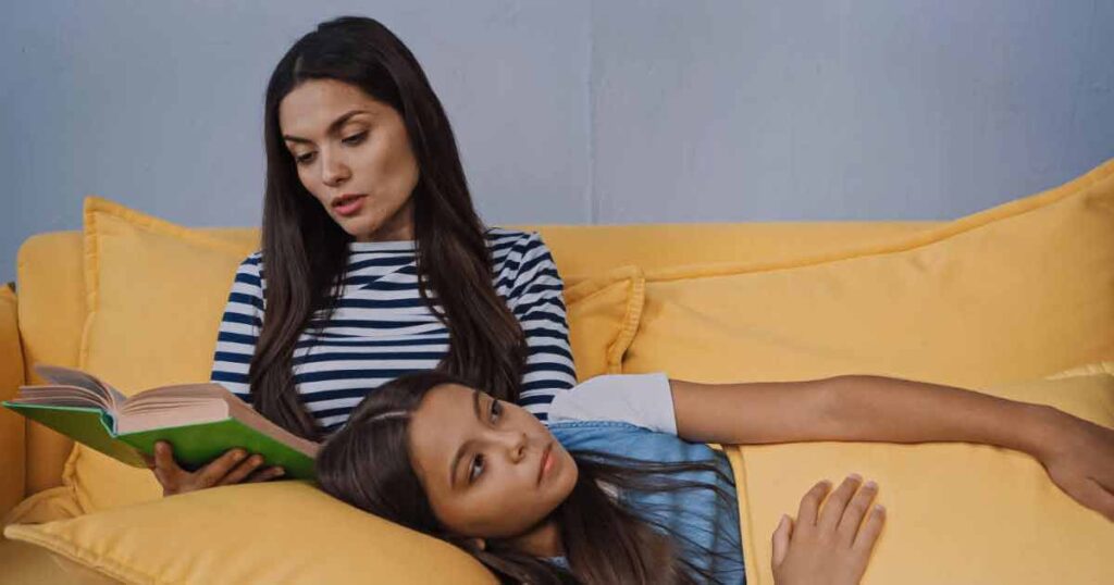 A woman sitting on a yellow couch reads a book aloud while a young girl lies beside her, resting her head on the woman's lap and listening attentively.