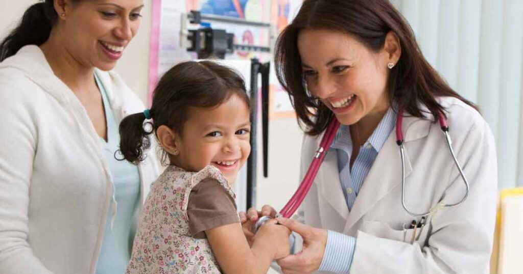 A smiling female doctor with a stethoscope examines a cheerful young girl, while a woman, likely her mother, stands beside them, also smiling, in a bright medical office.