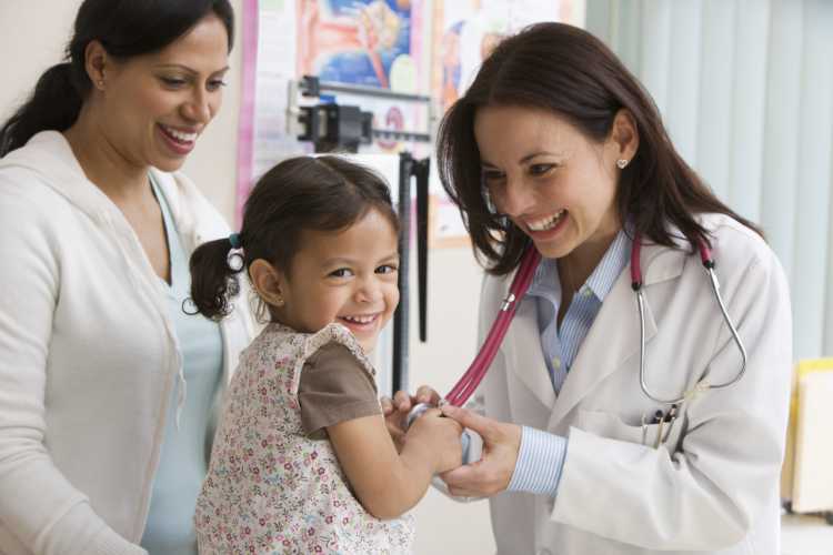 A smiling doctor with a stethoscope holds a little girl’s hand while she sits on an exam table, as her mother stands beside her, all appearing happy in a bright medical office.