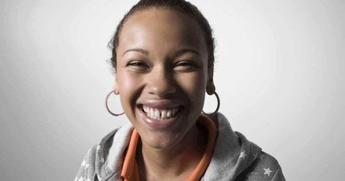 Una mujer joven con pendientes de aro sonríe alegremente a la cámara, vistiendo una sudadera gris con estrellas blancas y una camisa con cuello naranja, sobre un fondo claro y liso.