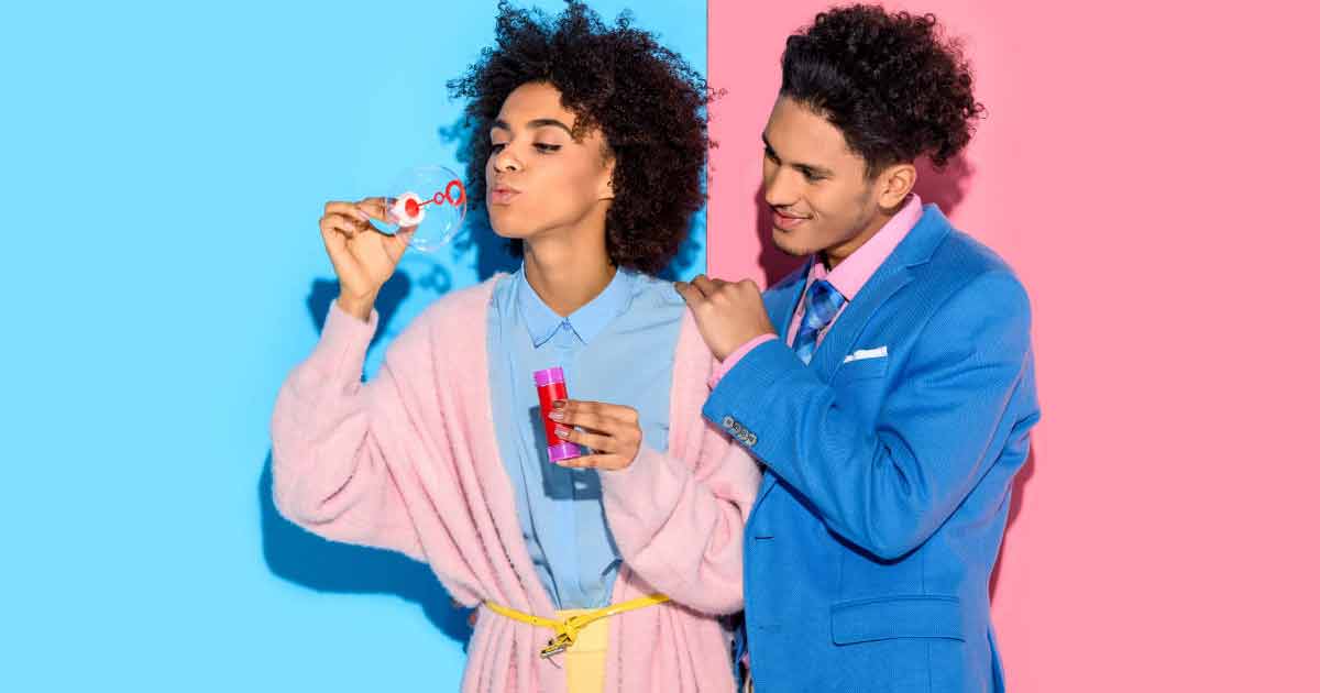 Una persona con un cárdigan rosa sopla burbujas mientras sostiene una pequeña caja de regalo, de pie junto a otra persona con un traje azul y una camisa rosa, sobre un fondo dividido en azul y rosa vibrante.