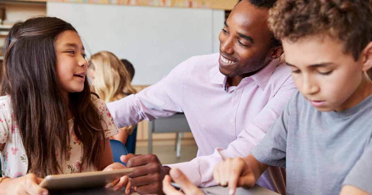 Una maestra sonríe mientras habla con una niña que sostiene una tableta, mientras un niño a su lado usa otro dispositivo en un salón de clases.