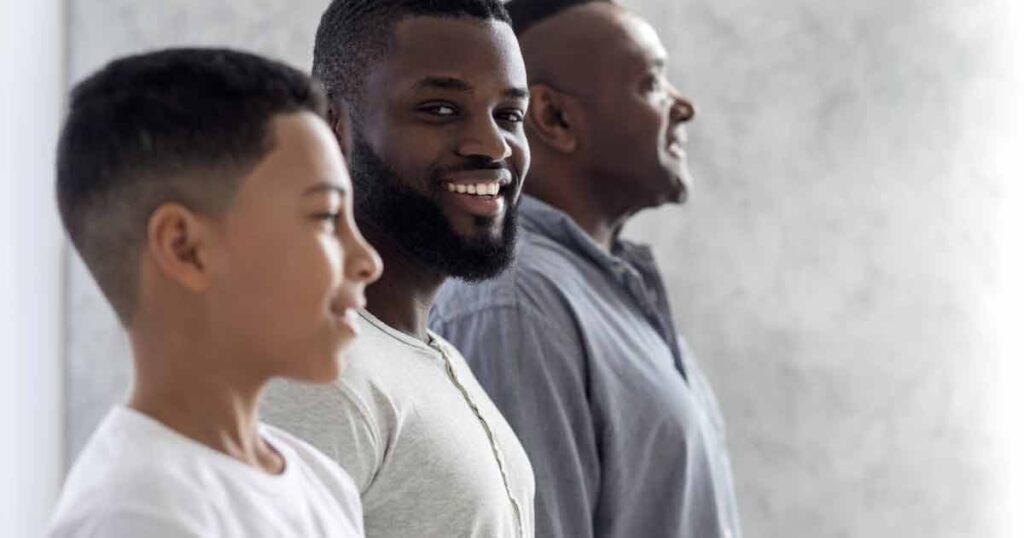 Three men of different ages stand side by side in profile, facing right. The youngest is in the foreground, an adult man is in the middle smiling, and an older man is in the background. The background is softly lit.
