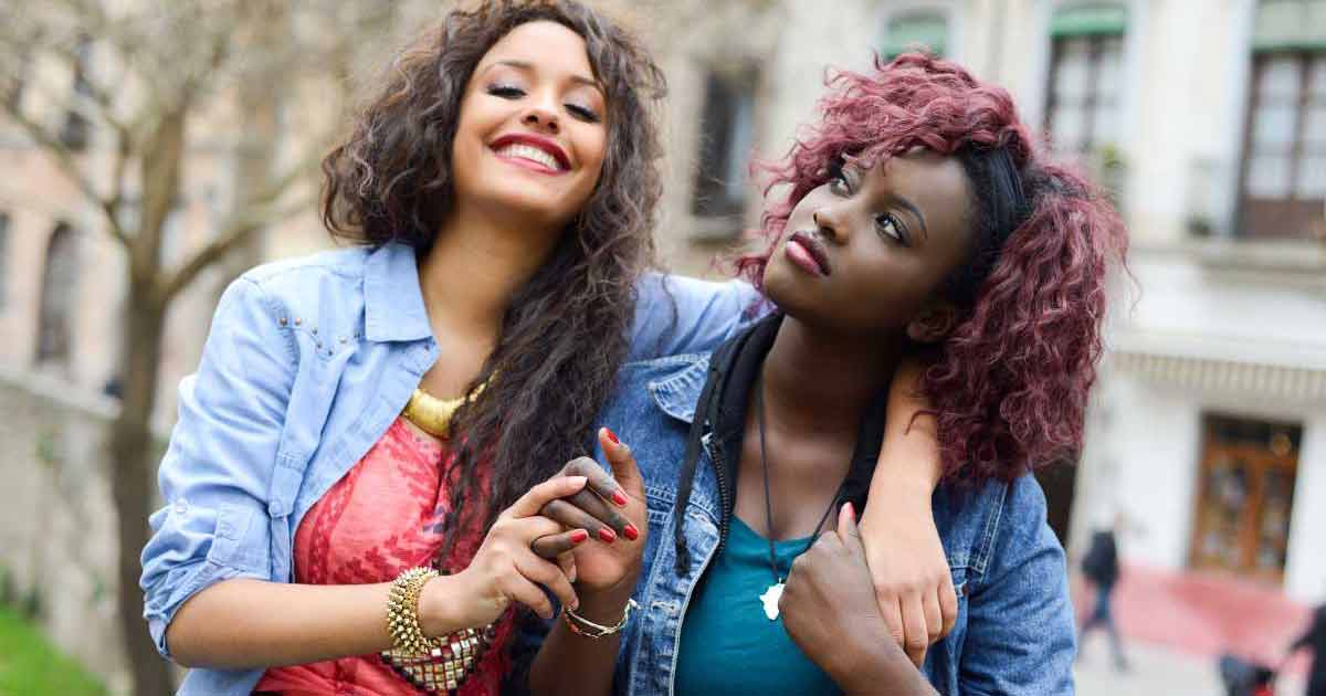 Two young women standing close together outdoors, smiling and holding hands. One woman has her arm around the other's shoulders, both wearing casual jackets and colorful tops, with a blurred background of buildings and trees.