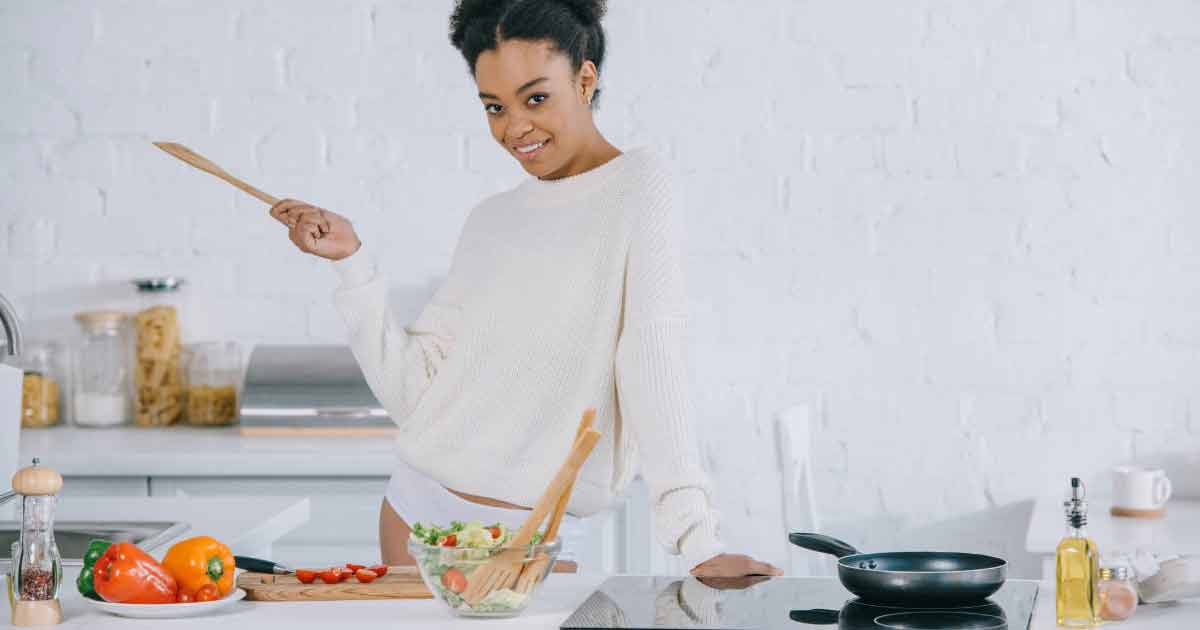 Una mujer con un suéter blanco está de pie en una cocina luminosa, sonriendo y sosteniendo una espátula de madera. En la encimera hay verduras frescas y una ensaladera, con una sartén cerca.