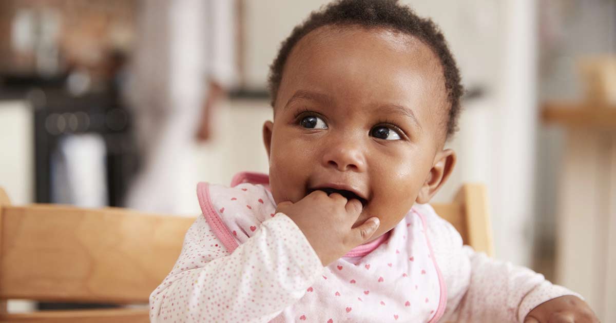 A baby wearing a white and pink outfit with a bib sits in a high chair, smiling and looking to the side with fingers in their mouth. The background is softly blurred, suggesting a home setting.