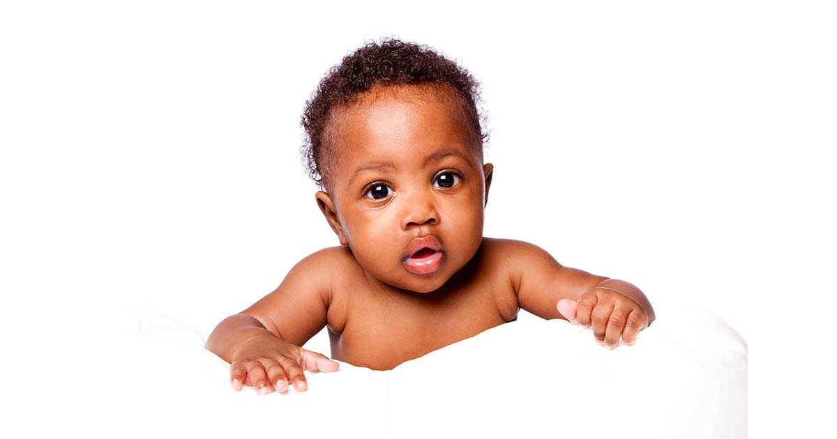 A baby with short curly hair looks forward with wide eyes and a slightly open mouth, resting both hands on a white surface against a plain white background.
