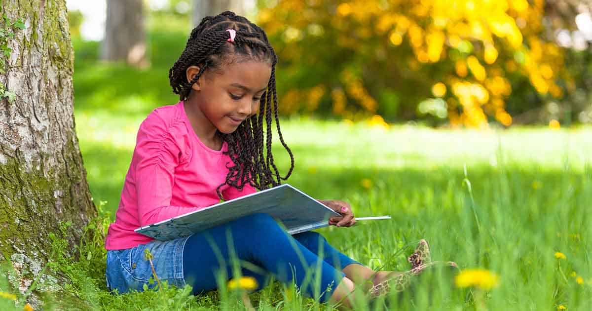 A young girl with braided hair sits on grass by a tree, reading a book. She wears a pink shirt, blue leggings, and leopard-print shoes. Bright yellow flowers and greenery are visible in the background.