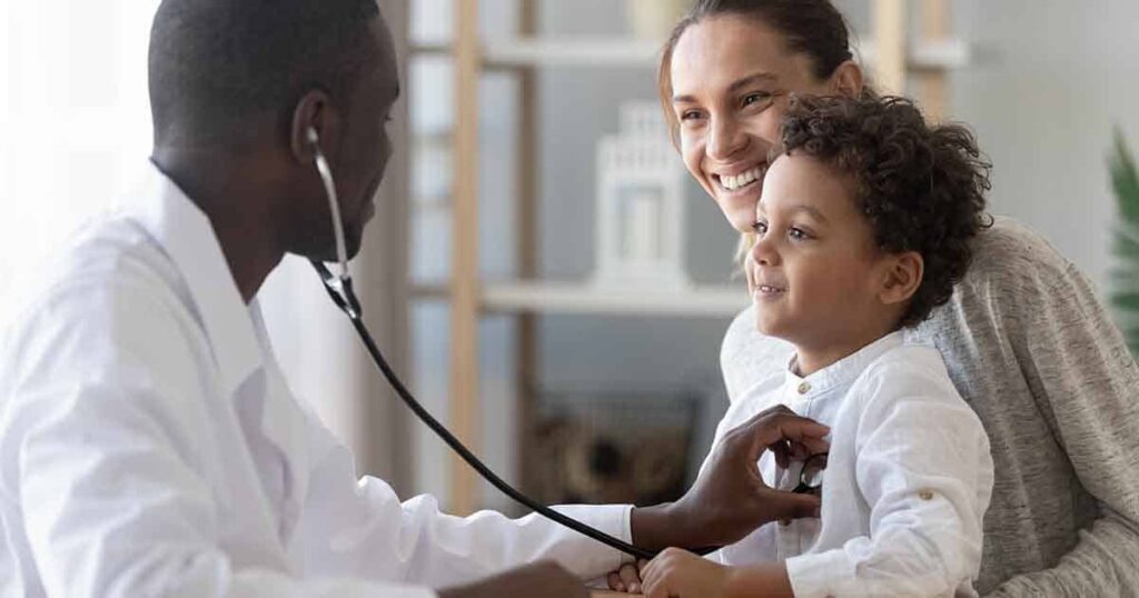 A doctor uses a stethoscope to listen to a young boy's chest as his smiling mother sits beside him in a bright room.