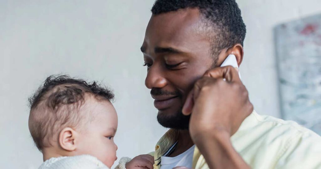 A man smiles while holding a baby in his arms and talking on a smartphone, standing indoors with a light background.