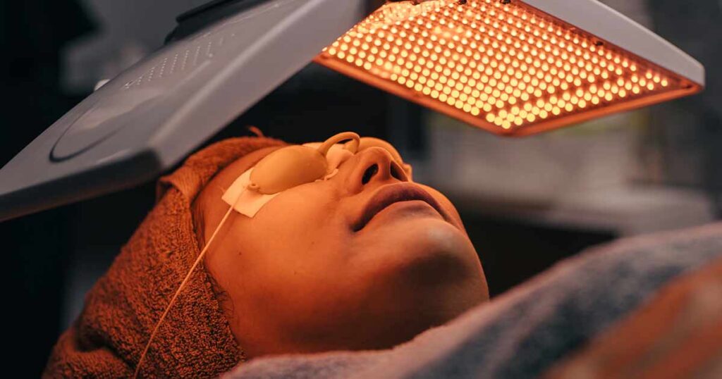 A person with a towel wrapped around their head lies under a panel of orange LED lights, wearing protective goggles over their eyes during a light therapy facial treatment.