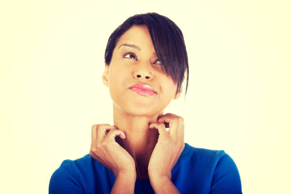 A woman wearing a blue top looks up thoughtfully while scratching her neck with both hands, lips pressed together, appearing contemplative or uncertain, against a light background.