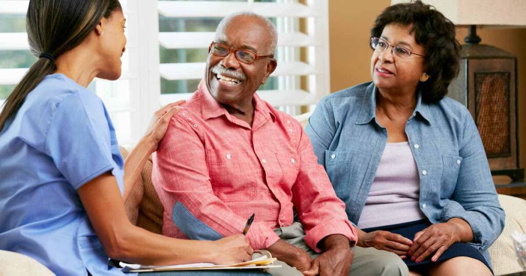 A nurse sits with an elderly couple on a couch, smiling and holding a clipboard while talking. The man and woman listen attentively, and the atmosphere appears friendly and supportive.