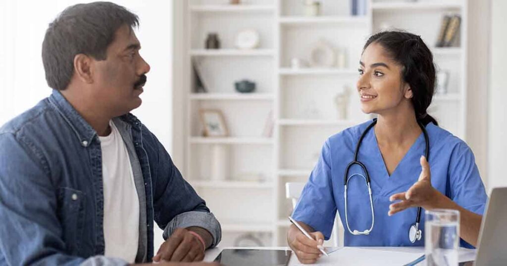 A healthcare professional in blue scrubs and a stethoscope talks with a seated male patient, gesturing and smiling as she takes notes during their consultation in a bright office.