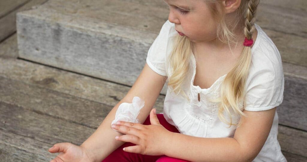 Young child with blonde hair sitting on wooden steps, wearing a white shirt and red pants, applying a white cream or lotion to her arm while looking at it.