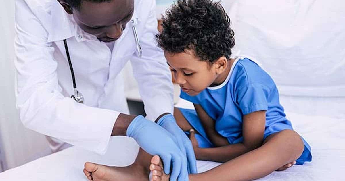 A doctor in a white coat and blue gloves examines the lower leg and foot of a young boy sitting on an exam table, who looks down at his leg with a concerned expression.